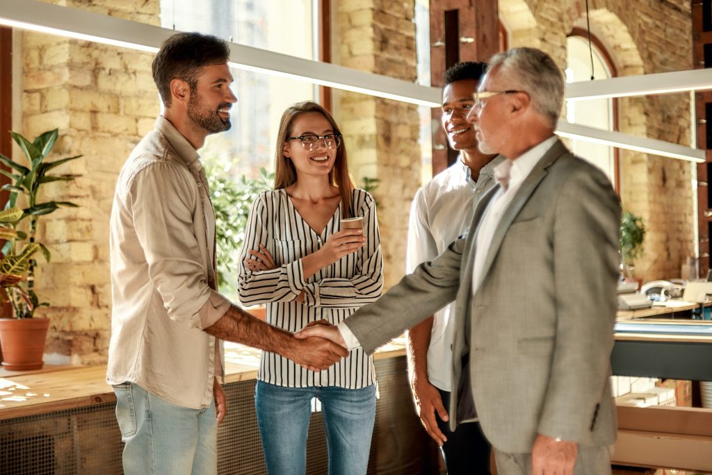 Two business people shaking hands and smiling while standing with coworkers in the creative office